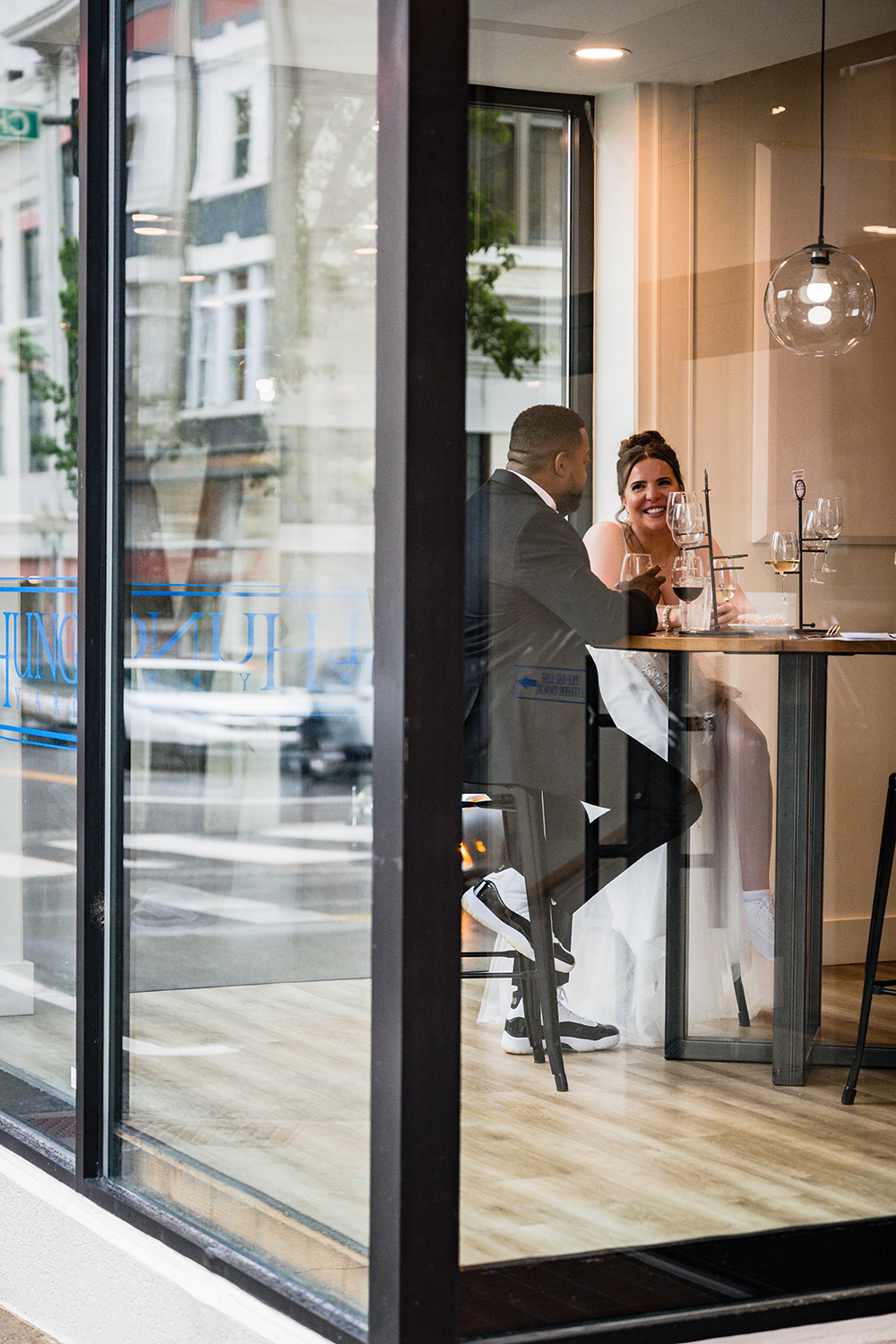 A couple is photographed through the glass outside of Well Hung Vineyard in Downtown Roanoke. Both people are wearing wedding attire and are seated at a round table with wine flights on the table from the restaurant. The groom looks at the bride who is smiling at him.