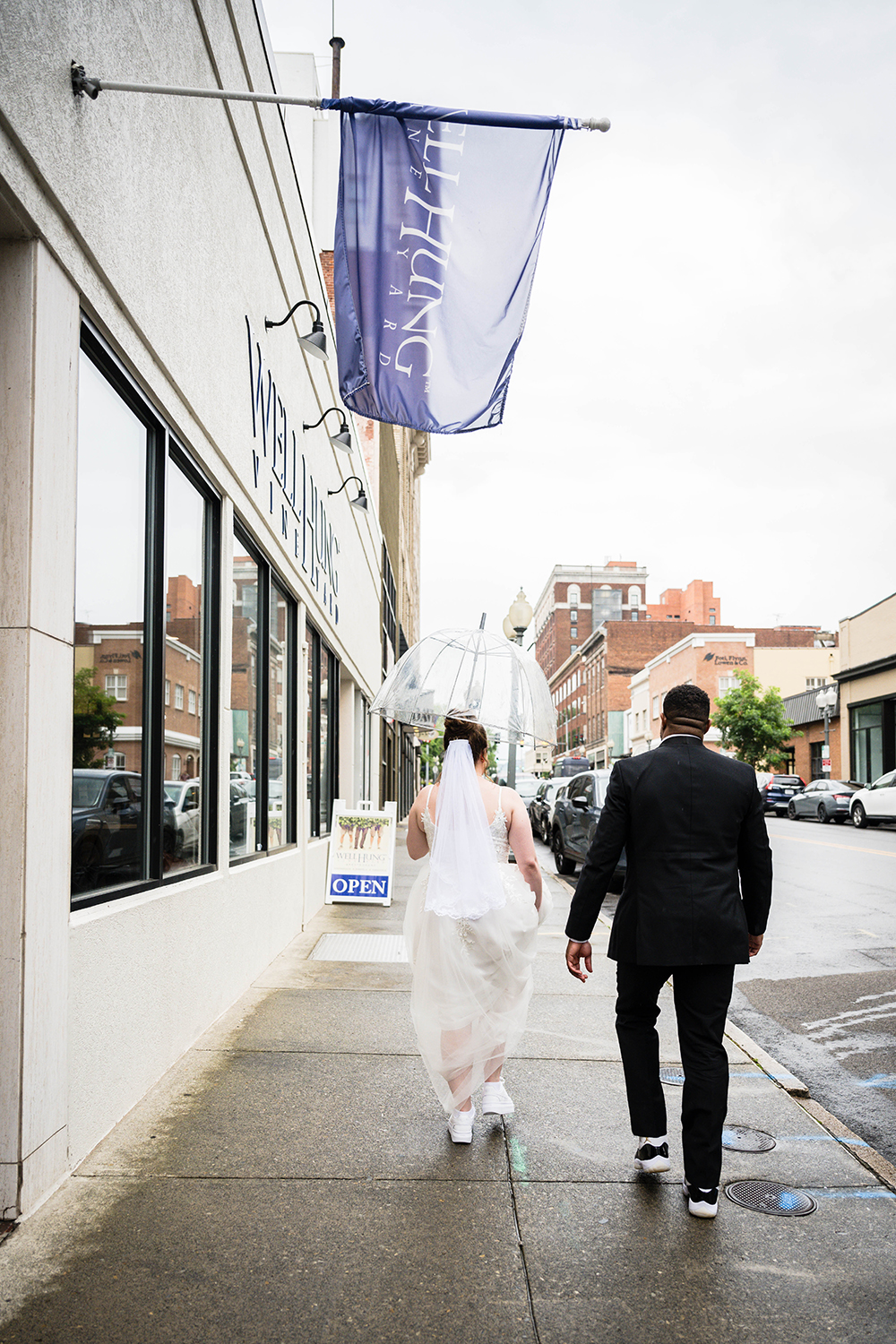 A bride holds a clear umbrella over her head and holds her dress as the groom follows closely behind. The pair are photographed from behind entering into Well Hung Vineyard in Roanoke, Virginia.