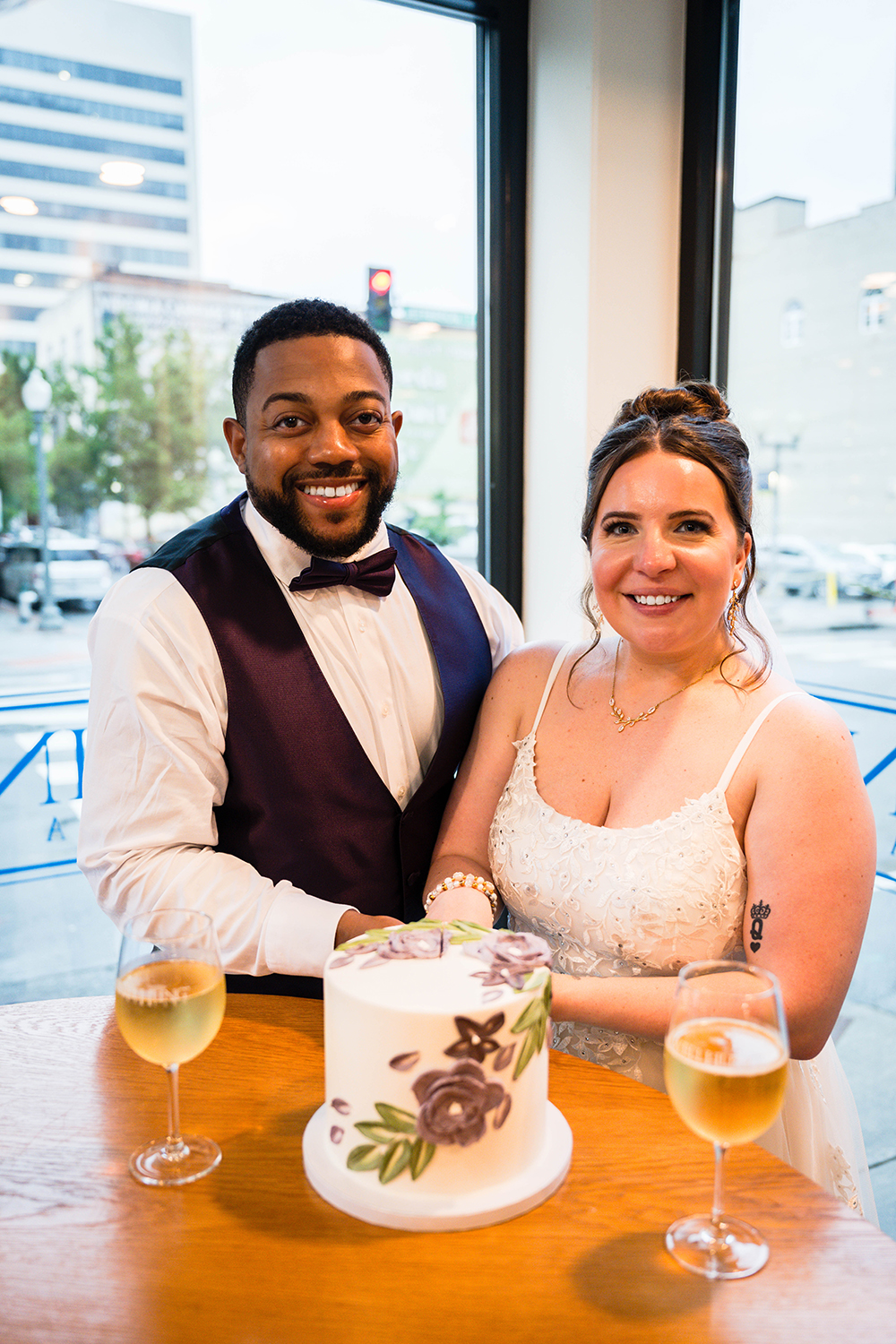 Two marriers cut into their cake and smile for the camera. They're inside Well Hung Vineyard, a restaurant in Downtown Roanoke in Southwest Virginia. The cake is being cut on a round table and is in the middle of two glasses filled with white wine.