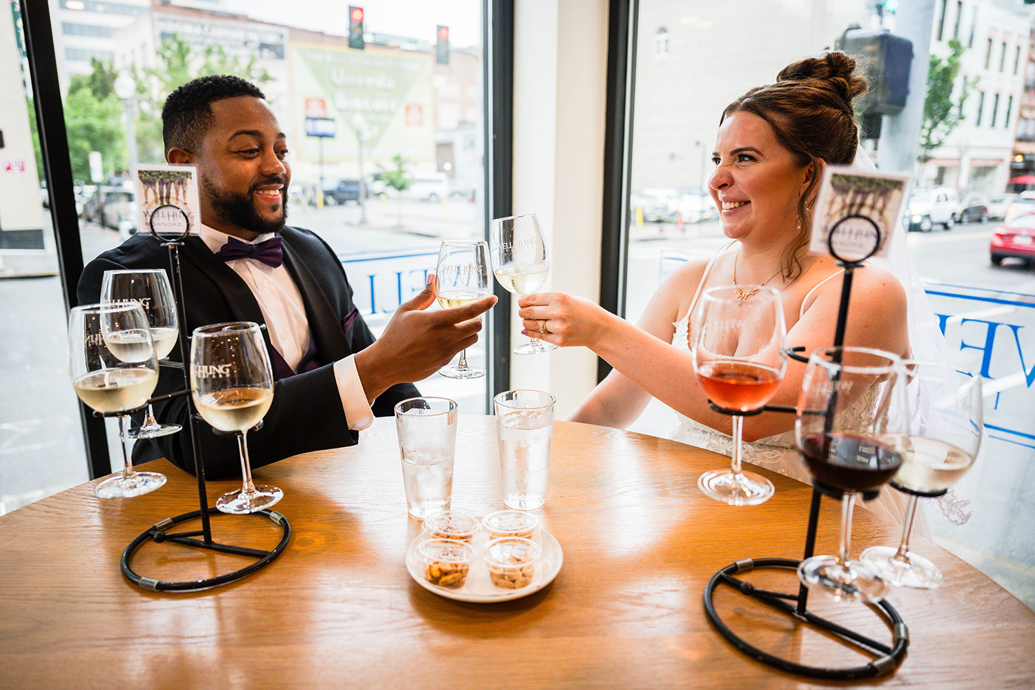 A couple sits at a round table in Well Hung Vineyard in Downtown Roanoke. The couple each grab their first wine glass from their wine tasting flight and clink their glasses while smiling at one another.