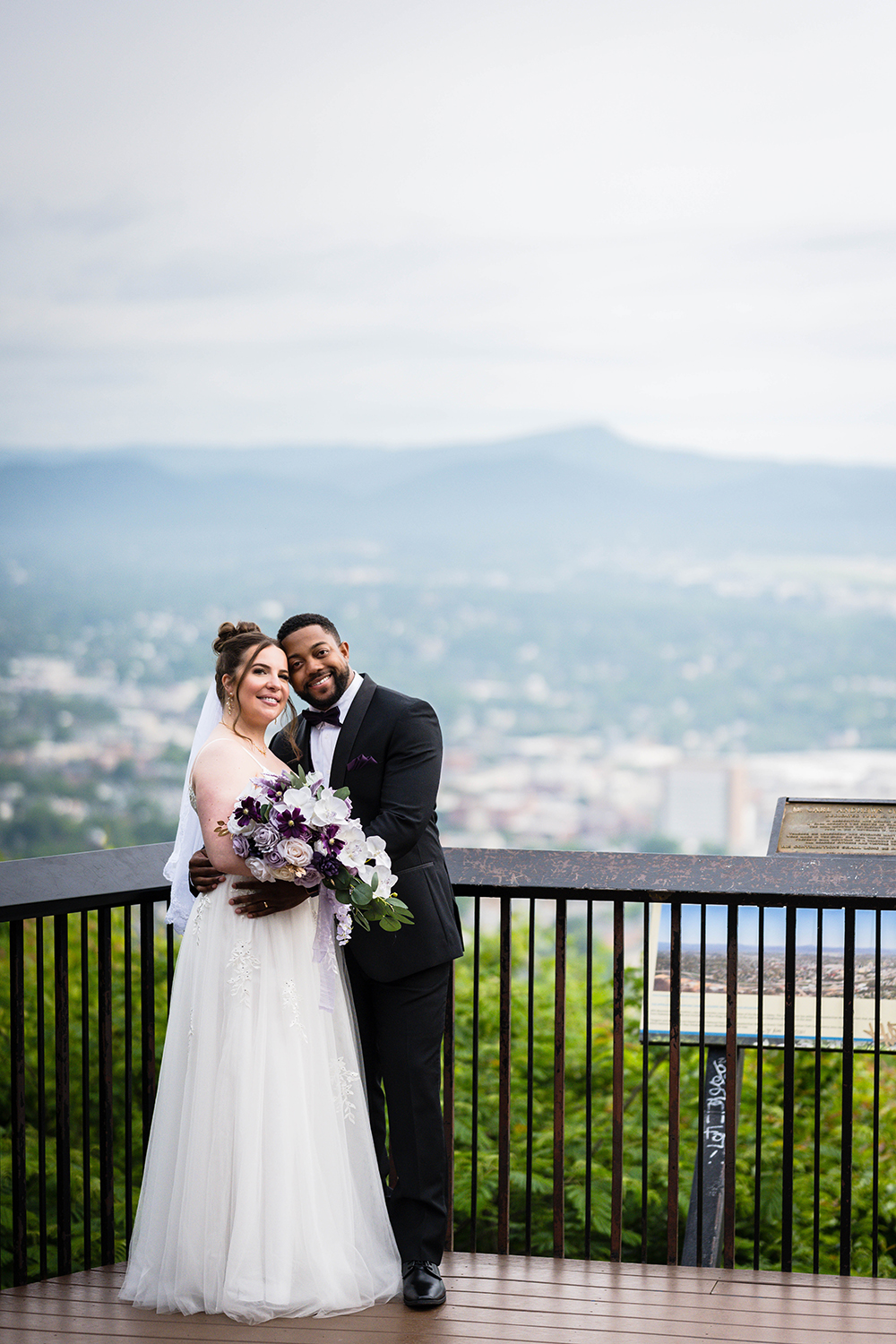 A couple embraces one another and smiles as they pose for a photo at the Roanoke Mountain Overlook, overseeing the Roanoke city skyline and the Blue Ridge Mountains.