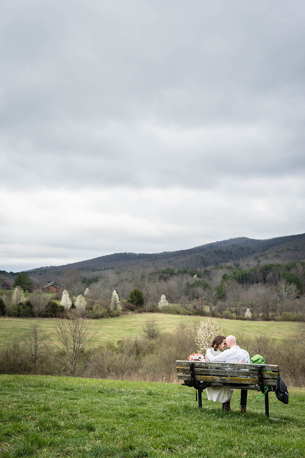 Two marriers sit on a bench at Heritage Park and press their foreheads on one another and smile widely. The view behind them is of the Blue Ridge Mountains on a cloudy day.