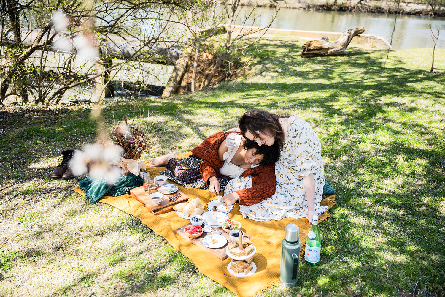 An LGBTQ+ couple snuggles together under a tree with speckled shade for a brunch picnic on their elopement day.