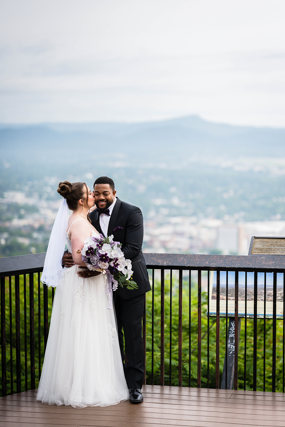 A bride gives a groom a kiss on the cheek as they stand at the Mill Mountain Overlook.