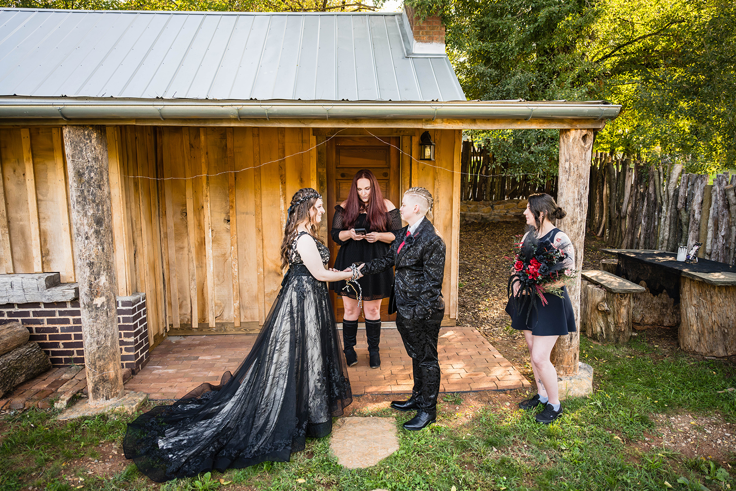 An LGBTQ+ couples performs a handfasting ceremony while their officiant reads from their phone and their witness holds one of the marrier's bouquet. The group stands outside on a small porch of an Airbnb,