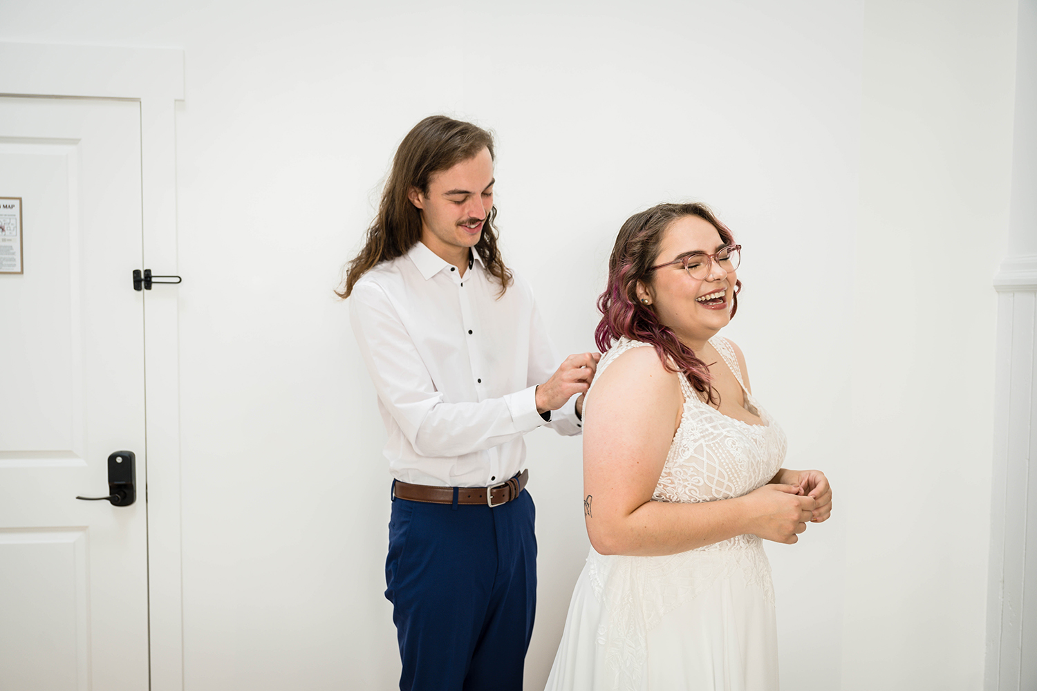 A partner helps his significant other button up her dress on their elopement day in their hotel room.