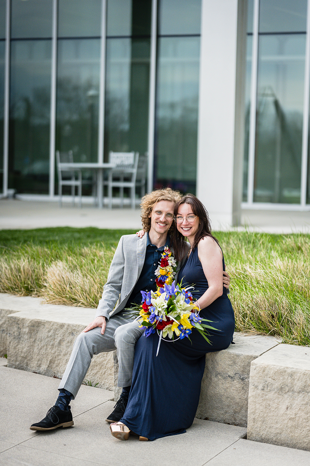 A couple on their elopement day sit on a cement ledge and wrap their arms around one another as they smile for a photo outside of the Moss Arts Center on Virginia Tech's campus. The person on the right places a wedding bouquet in her lap while the other person just leaves their hand on his leg. 