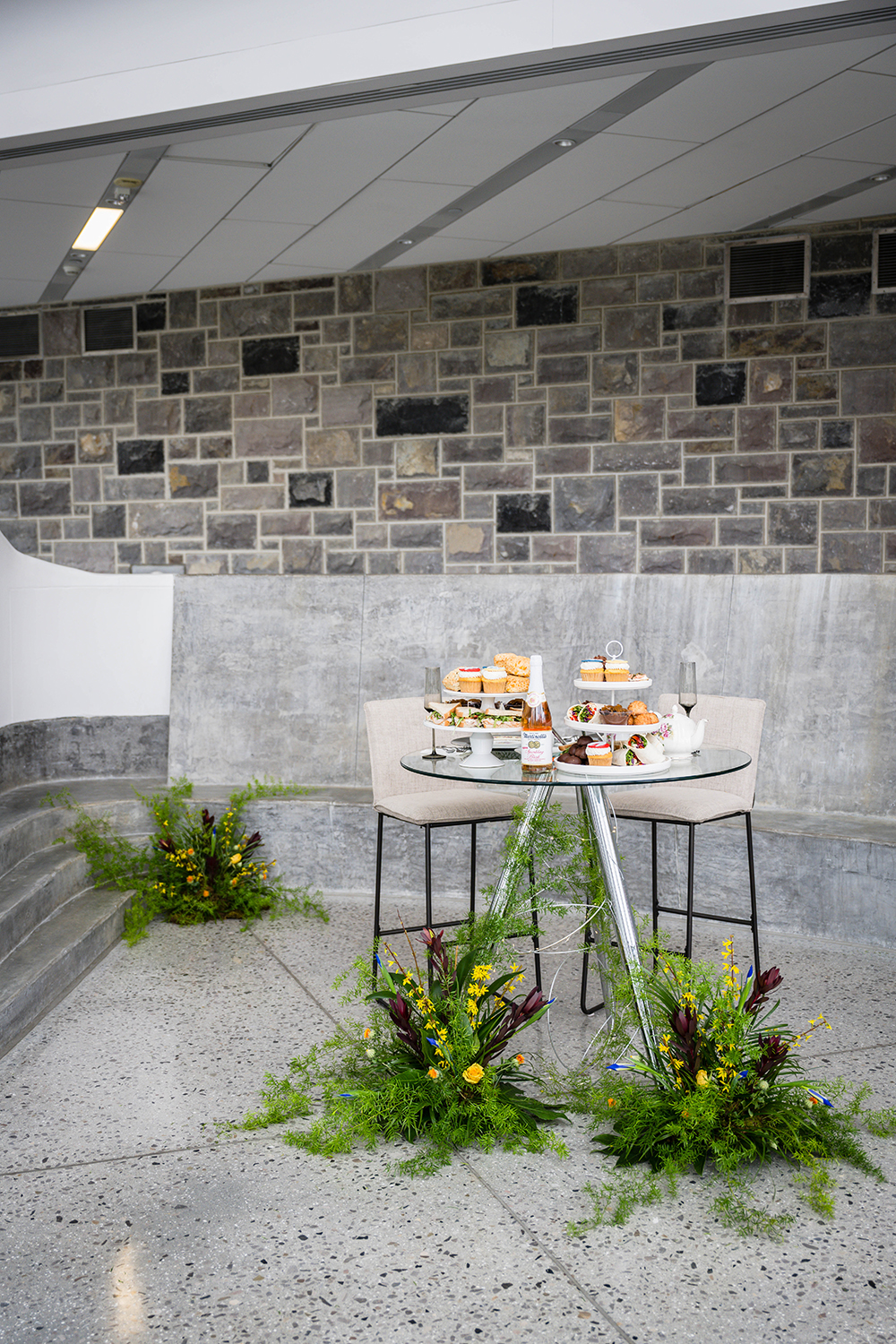 A clear circular table with silver legs is decorated with unique floral arrangements made by florists in Roanoke. On top of the table is two three-tired trays with tons of snacks, two champagne flutes, a bottle of non-alcoholic cider, and a teapot. Behind the glass table are two tall chairs. This table set up is located inside of the Moss Arts Center. 