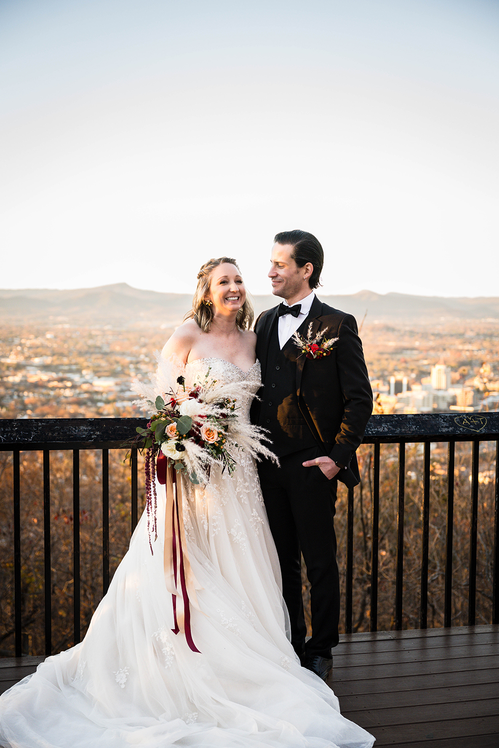 A bride and groom stand side-by-side on the Rockledge Overlook at Mill Mountain which oversees the Blue Ridge Mountains and the Downtown Roanoke city skyline as the sun begins to set. The groom angles himself towards his bride and smiles softly as she lets out a large grin. She is holding a big and imposing bouquet with strands of ribbon dangling by her side.