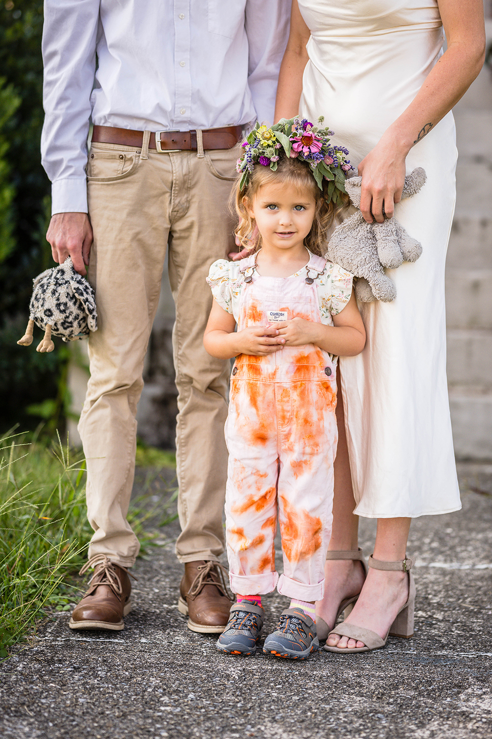 A child stands directly in front of their parents (who you can only see from their chest down) and looks shyly at the camera for a photo. The parents are each holding onto one of the child's stuffed animals in their hands.