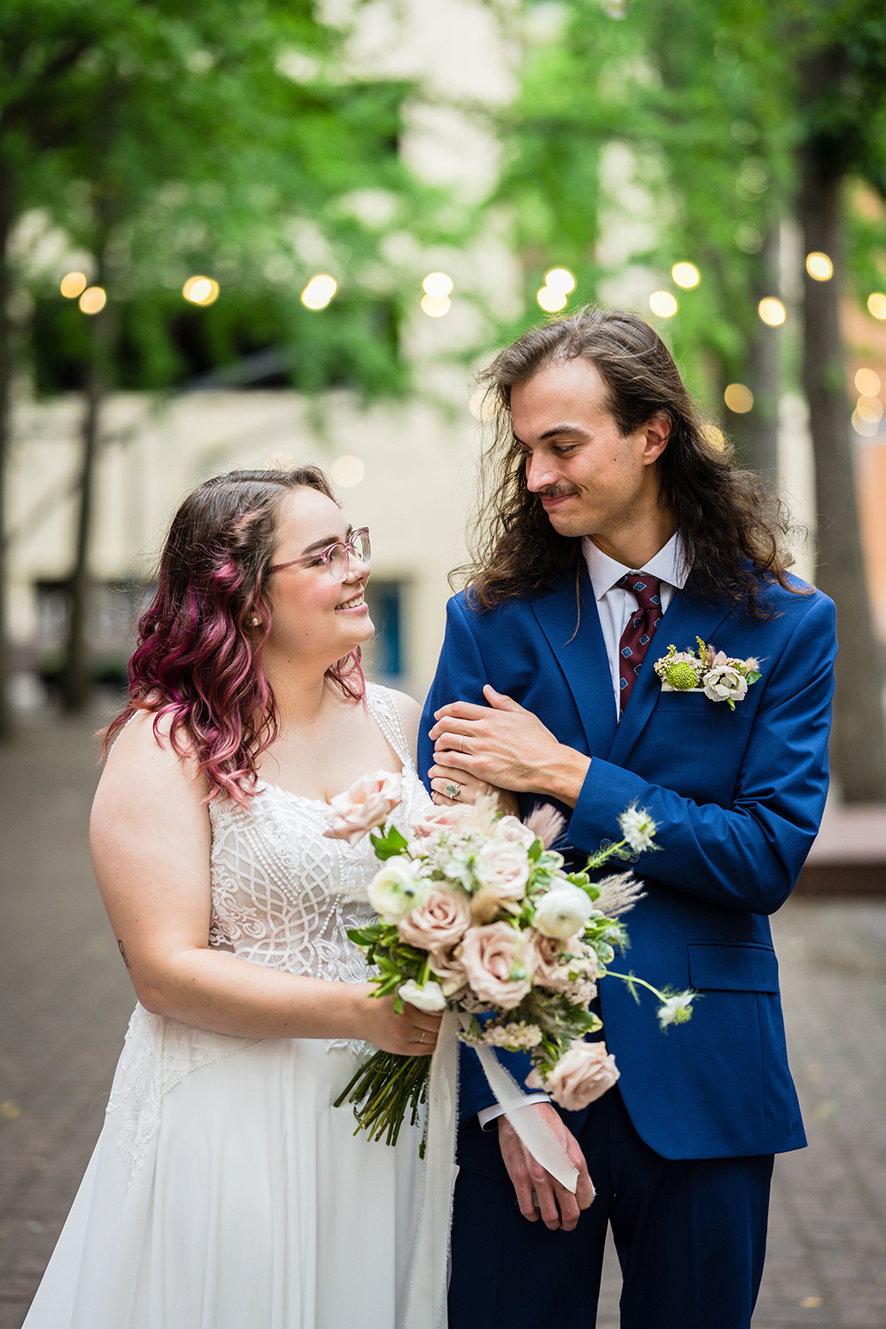 A couple looks and smiles at one another in Century Plaza in Downtown Roanoke. A marrier wearing a wedding dress extends her bouquet out in between her and partner. With her free hand, she wraps it around her partner's shoulder as her partner lays a hand gently on her hand resting on his bicep.