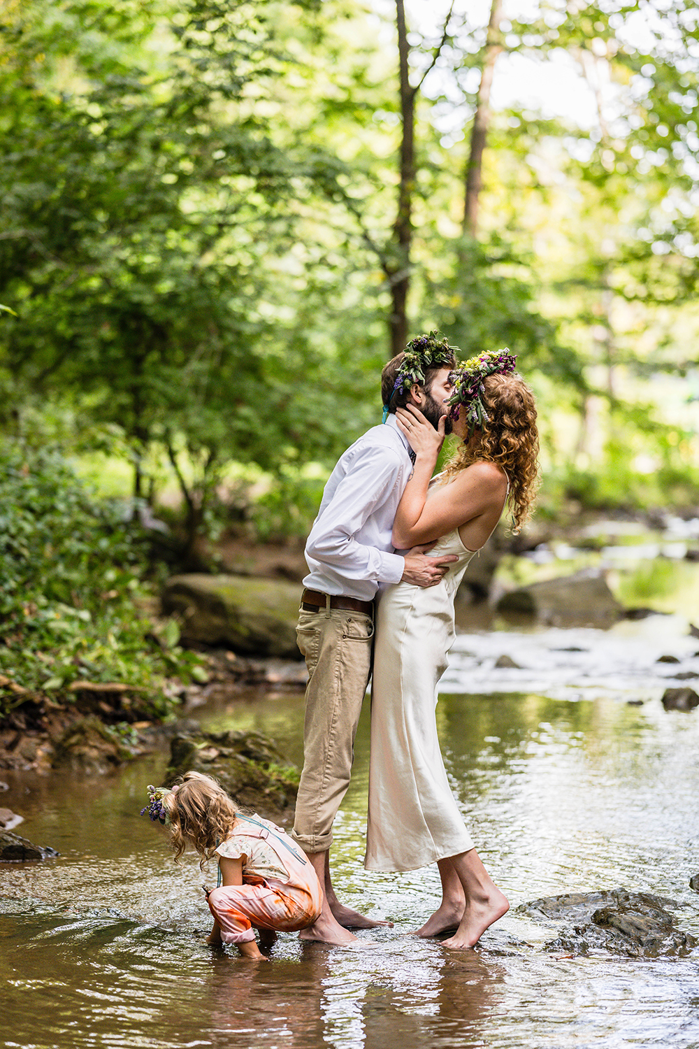 A couple go in for a kiss on their elopement day while their child squats to pick up a rock in front of them. All three of them are in the water at Fishburn Park in Roanoke, Virginia.