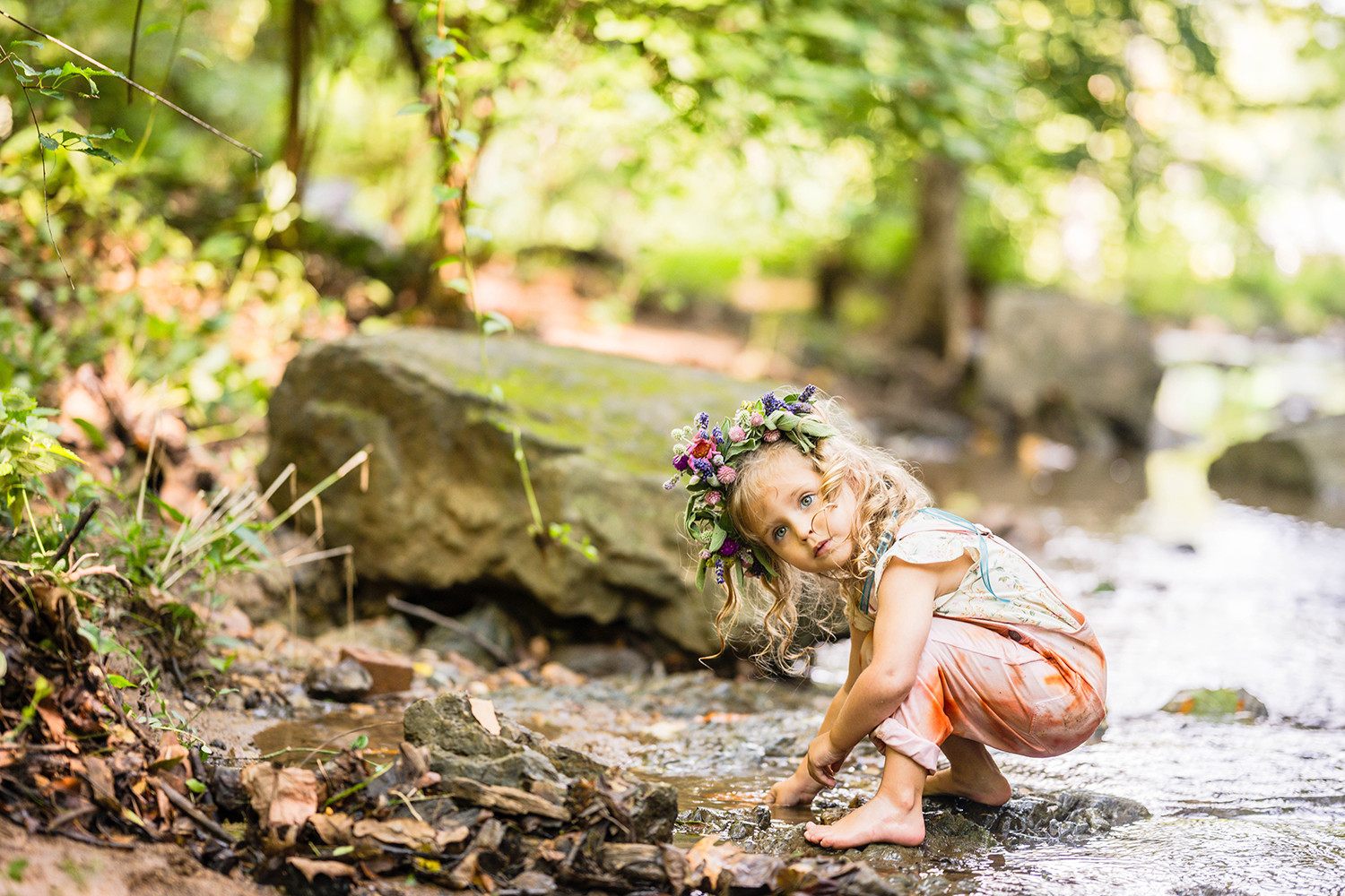 A child wearing a flower and eco-dyed suspenders plays in the water at Fishburn Park and momentarily pauses to look up and away from the camera.