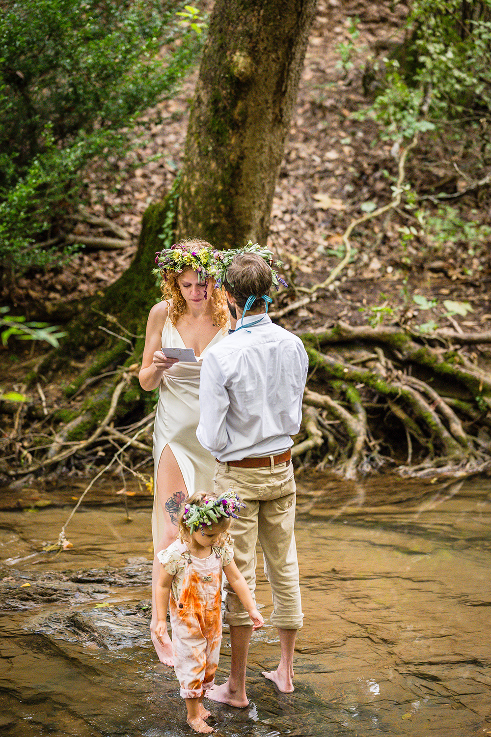 A couple reads their vows as they stand barefoot in the water at Fishburn Park on their elopement day. Their child walks in the water near them.