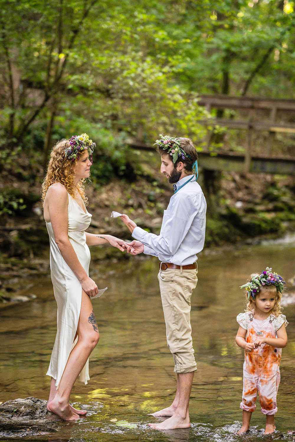 A couple reads their vows as they stand barefoot in the water at Fishburn Park on their elopement day. Their child walks in the water near them.