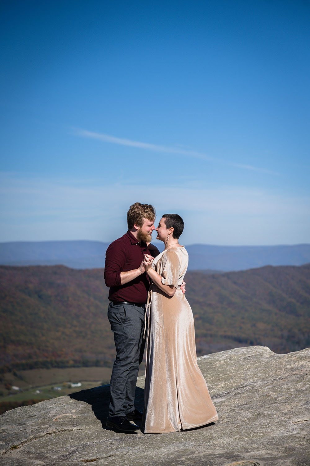 A couple holds onto one another and goes in for a kiss at a summit of a hike in the Blue Ridge Mountains. 