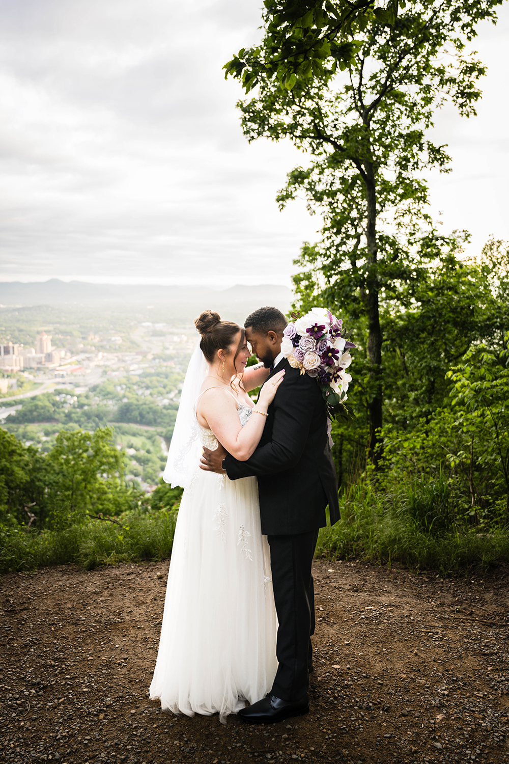 A couple hold one another and look at one another with the Roanoke city skyline and the mountains surrounding the city in the background.