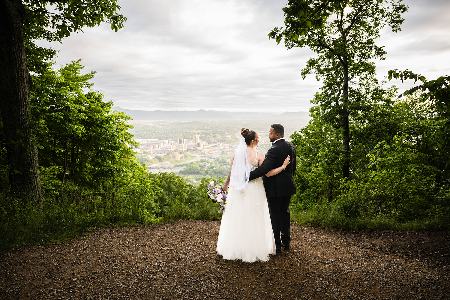 A couple on their elopement day in Roanoke, VA wraps their arms around one another and looks at each other as they enjoy the view of the Roanoke city skyline and the surrounding mountains.