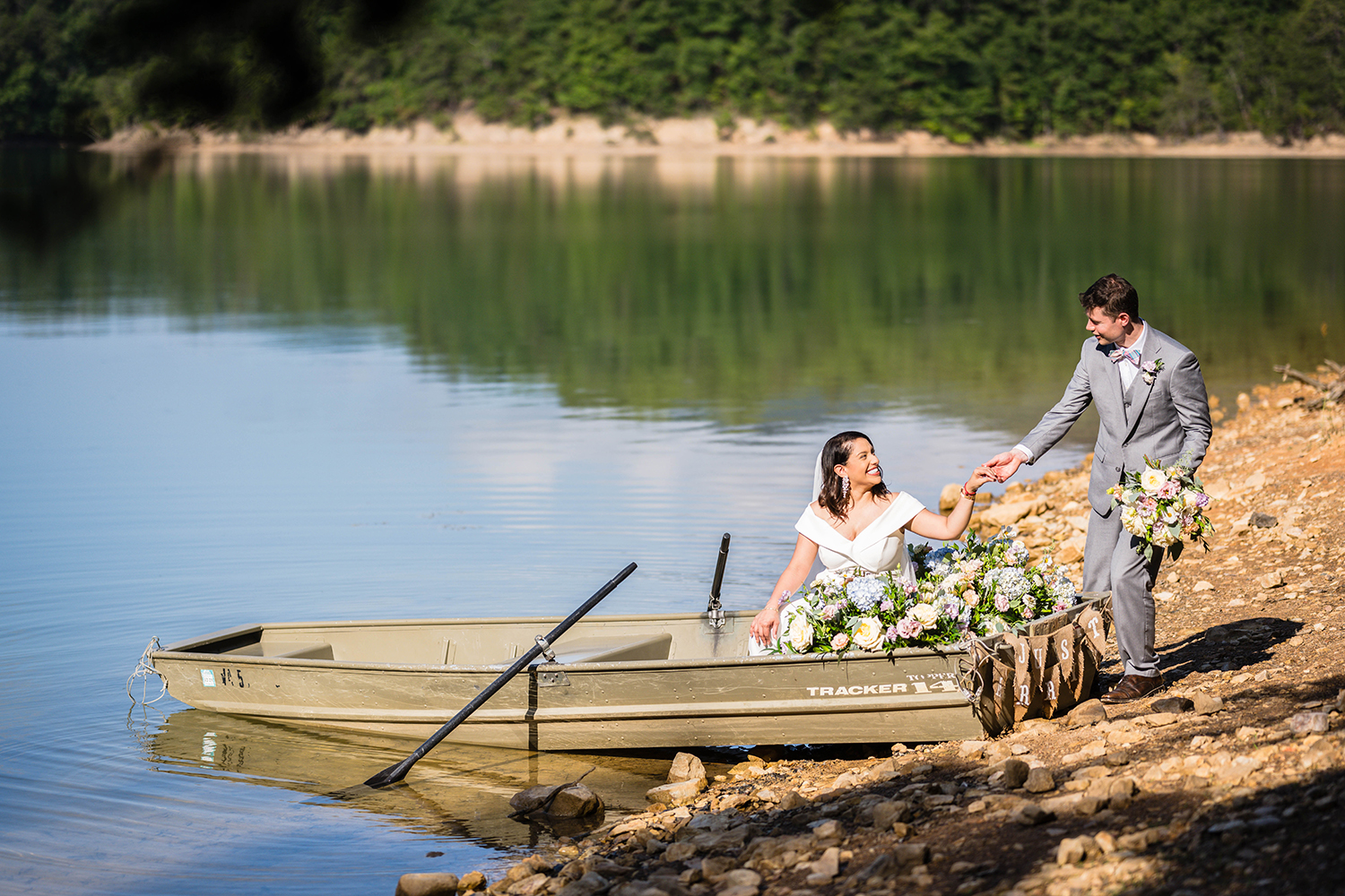 A partner helps his significant other into a rowboat adorned with flowers along the coast of Carvin's Cove on their elopement day while holding his significant other's bouquet. The significant other, already seated in the boat, keeps her hand in her partner's hand and looks up at him with a smile. 