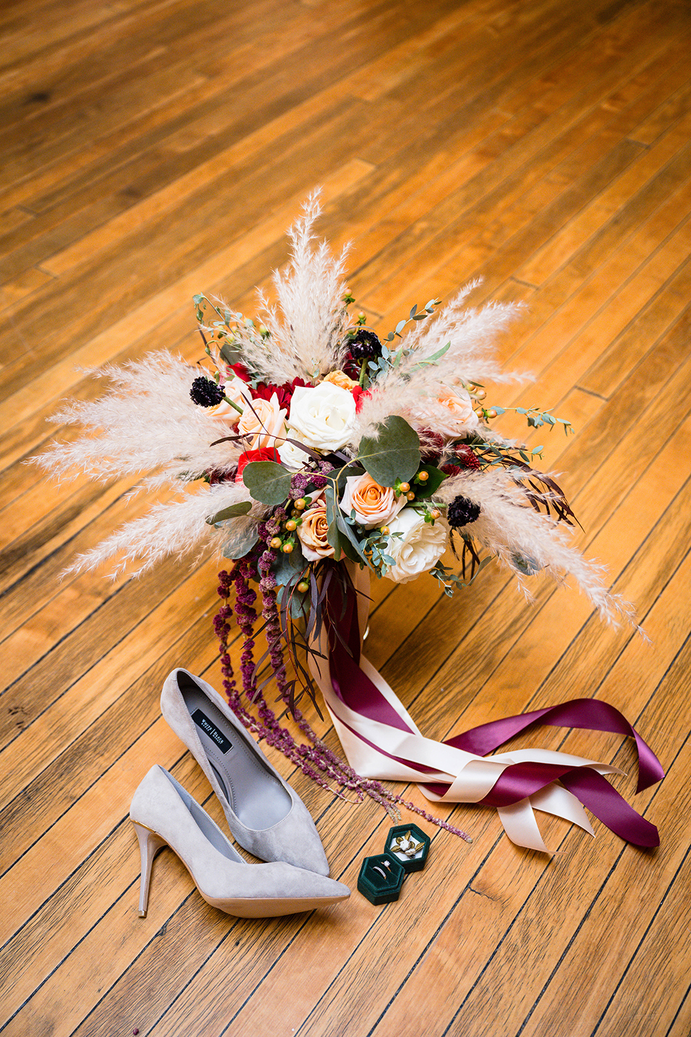 A bouquet of flowers in a glass vase is set on the floor next to the marrier's heels and ring box, which features her earrings and wedding ring. The bouquet's long flowing ribbons cascade down the vase and are neatly arranged along the floor.