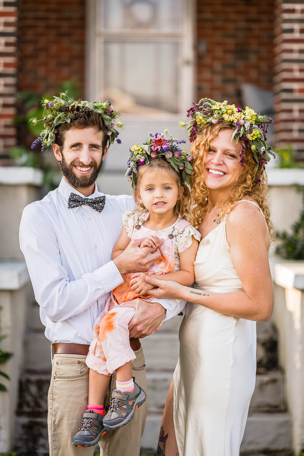 A family of three huddles together and smiles outside of their home. The trio are adorned with flower crowns. 