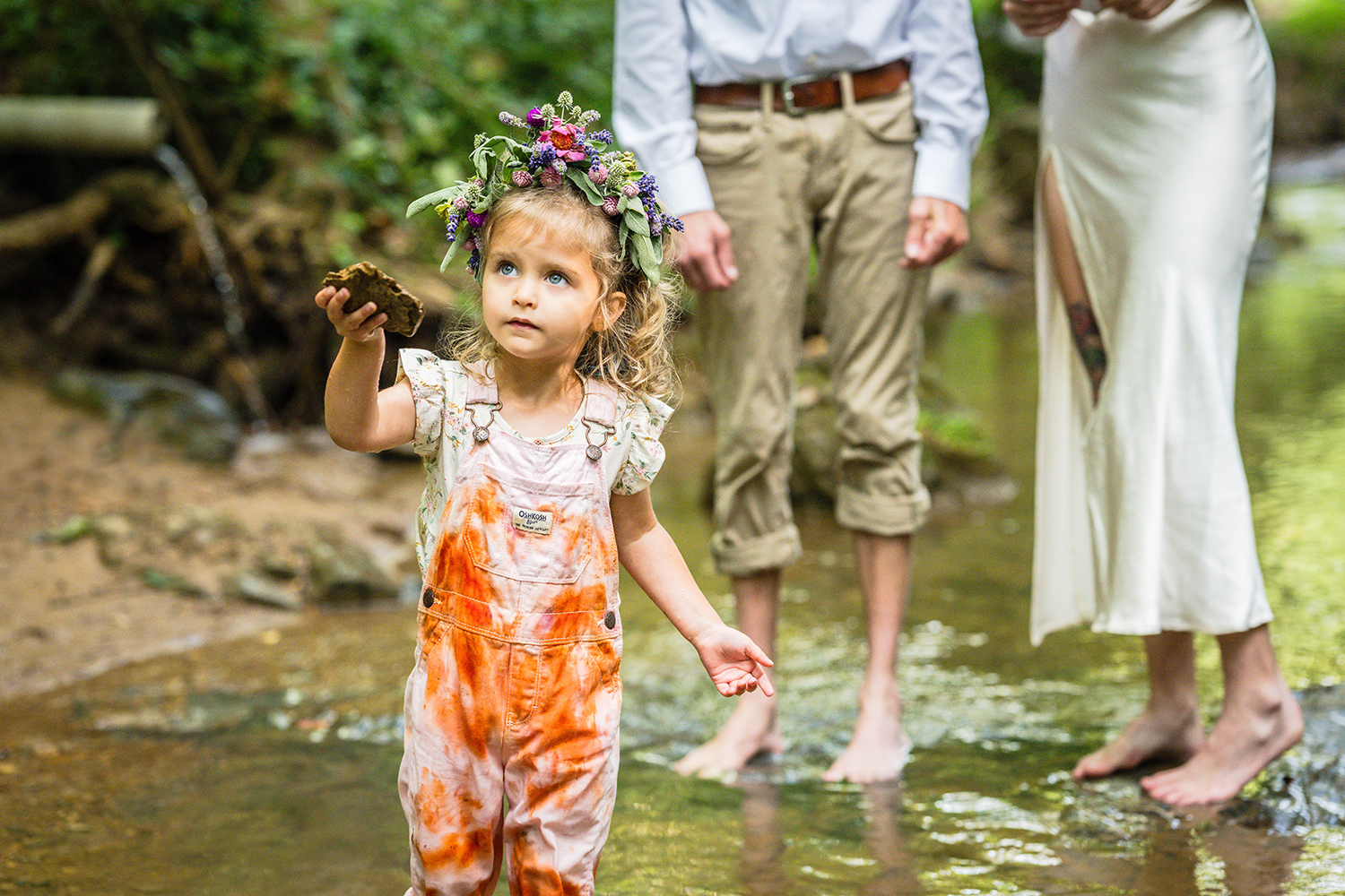 A child wearing a flower crown extends a flat rock out to the photographer (not seen in image) and looks slightly past the camera. The child's parents are seen in the background, but only from the waist down. The three of them are standing in a shallow creek at Fishburn Park.