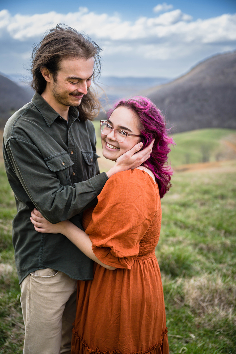 A woman looks at the camera smiling with her arms touching her partner's stomach as her partner holds her face and smiles widely during their anniversary adventure photoshoot in Giles County, Virginia.