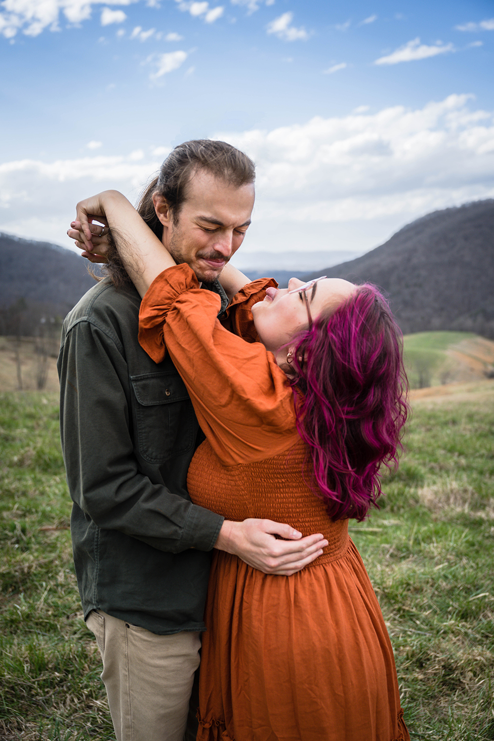 A woman wraps her arms around her partner's neck and sticks a tongue out at him while her partner closes his eyes and jokingly winces. 