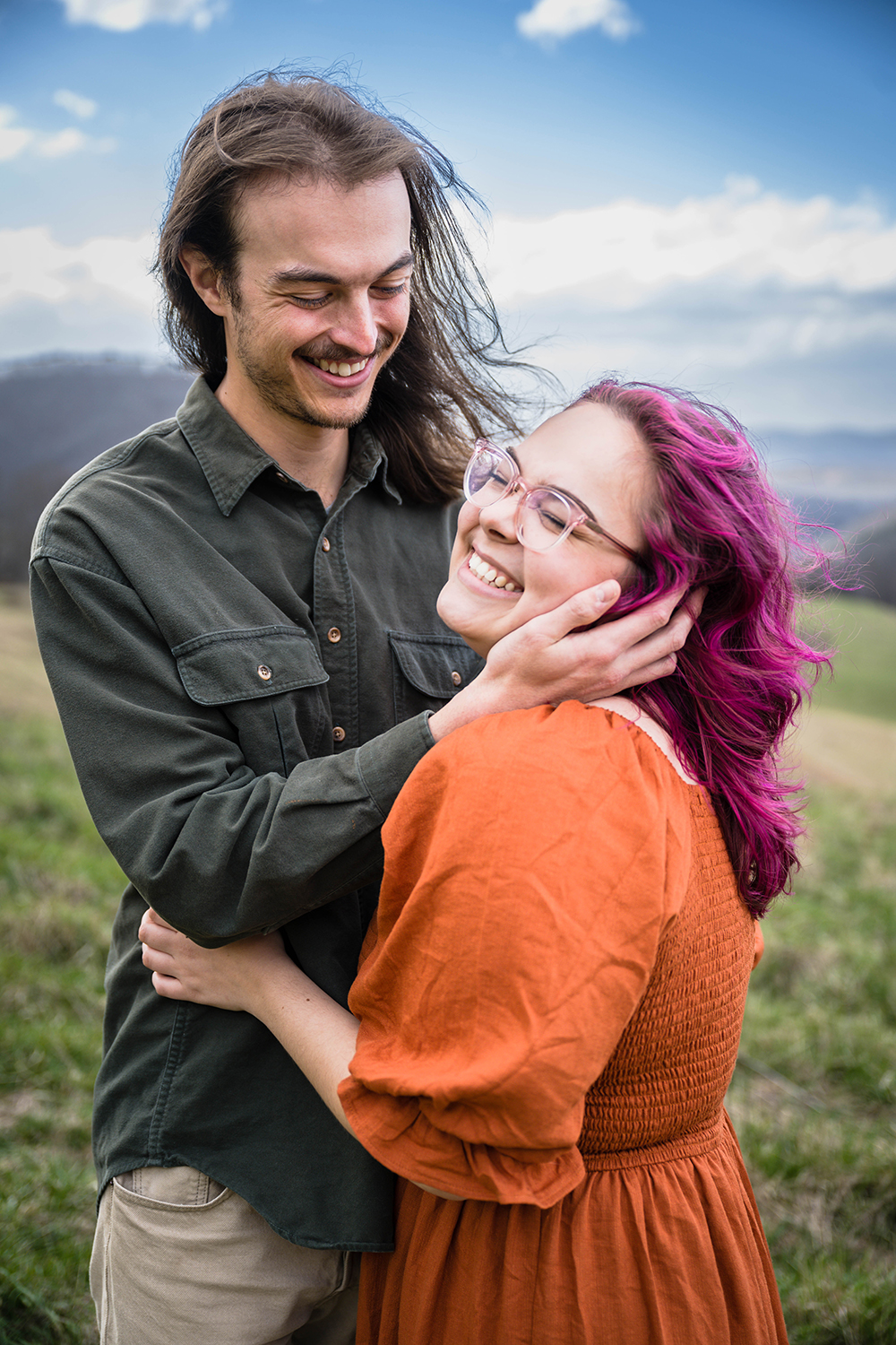 A woman smiles widely and closes her eyes tightly with her arms touching her partner's stomach as her partner holds her face and smiles.