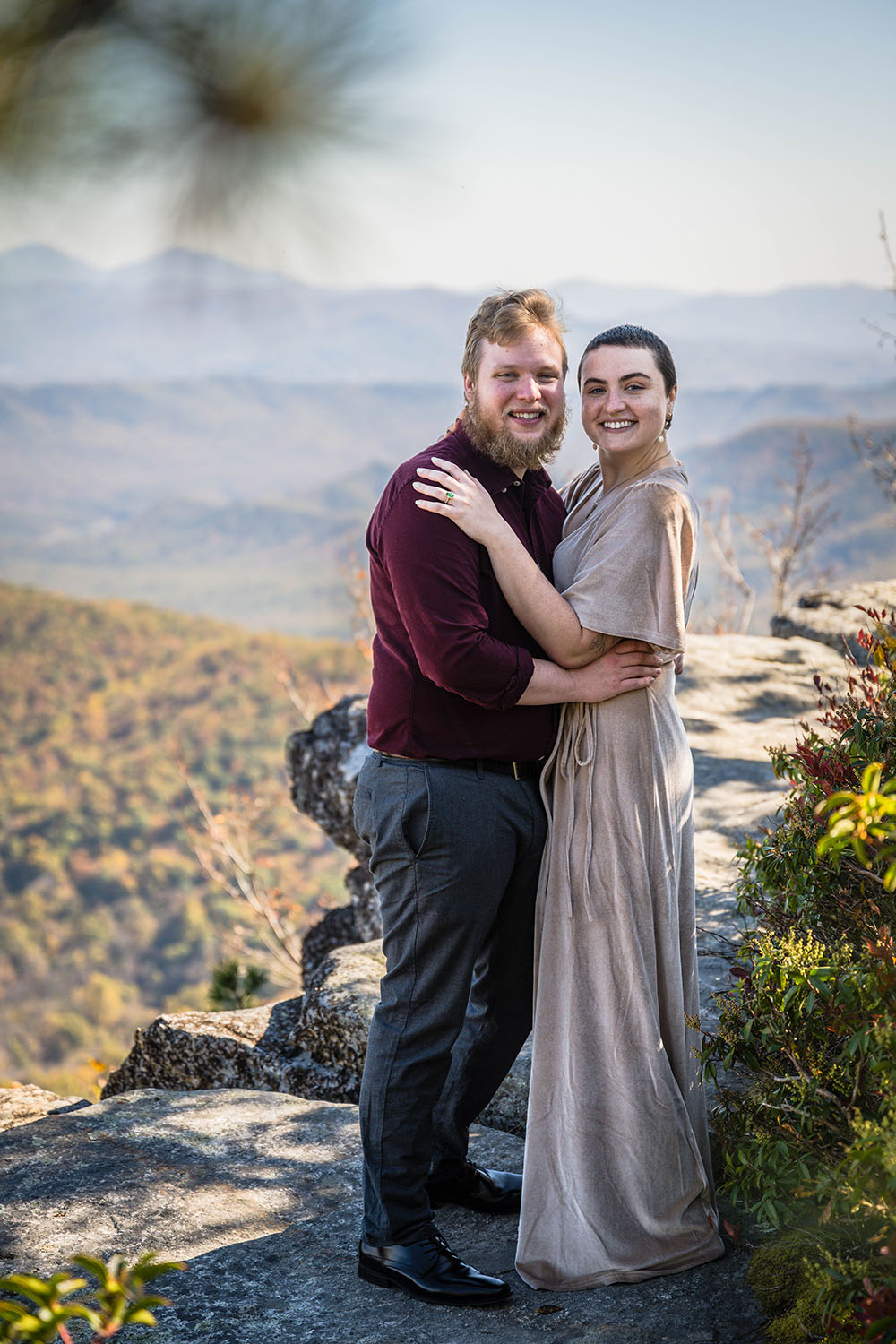 A couple wraps their arms around one another and smiles towards the camera as they stand on a rocky ledge with the Blue Ridge Mountains in the background.