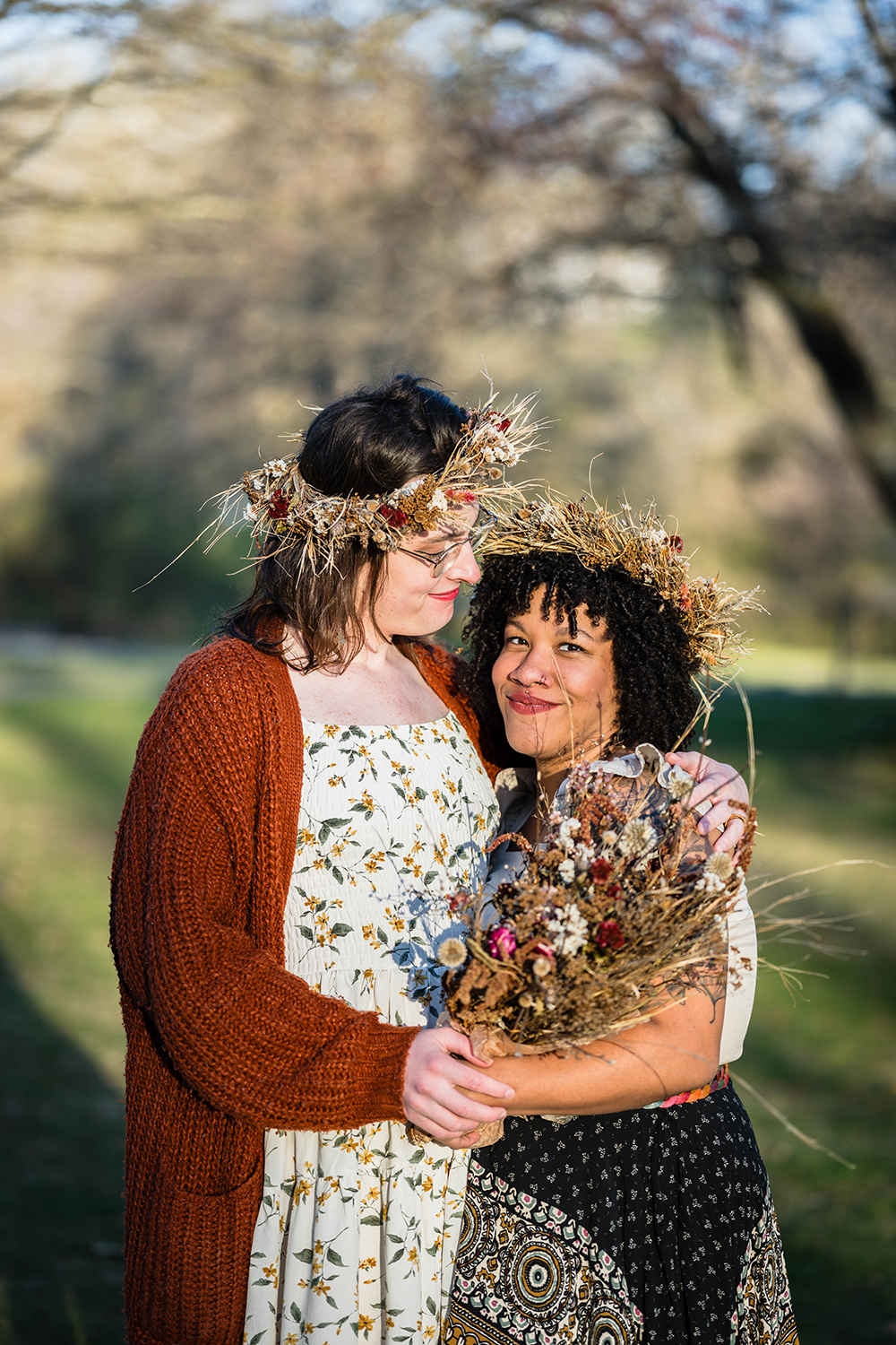 An LGBTQ+ couple wearing dried flower crowns stands together for a photo. A transwoman marrier looks down at her partner and smiles as her partner holds onto a large bouquet of dried flowers. 