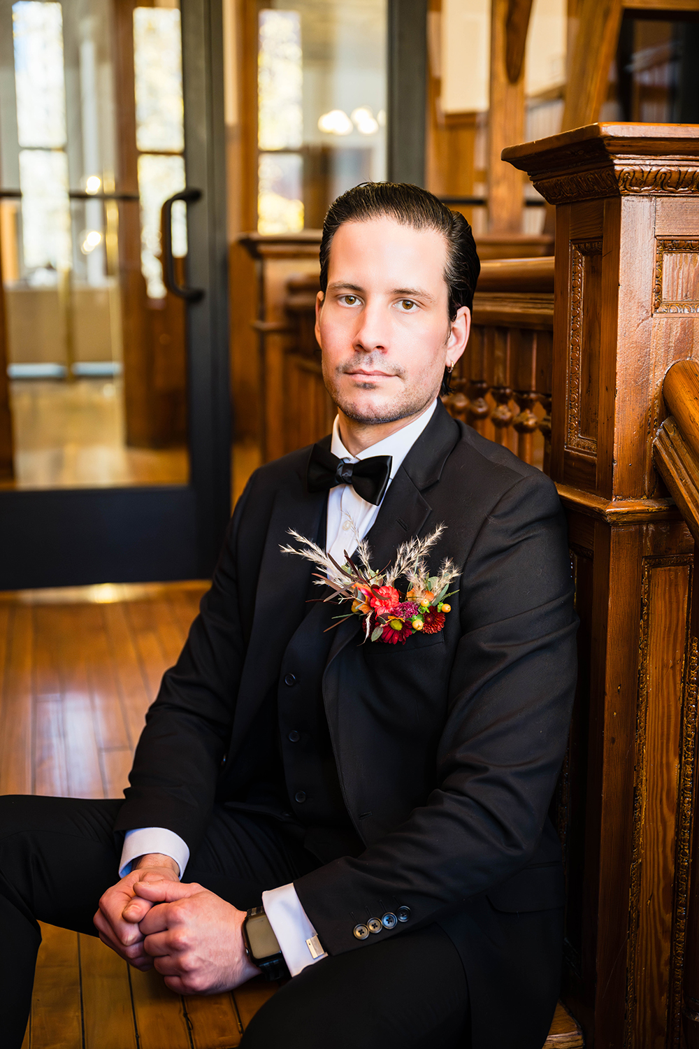 A groom sits at the edge of one of the first stairs in the stairwell of the Fire Station One Boutique Hotel in Downtown Roanoke. He has a serious expression on his face as she angles himself toward the camera to show off his floral pocket square arrangement in his suit jacket pocket.