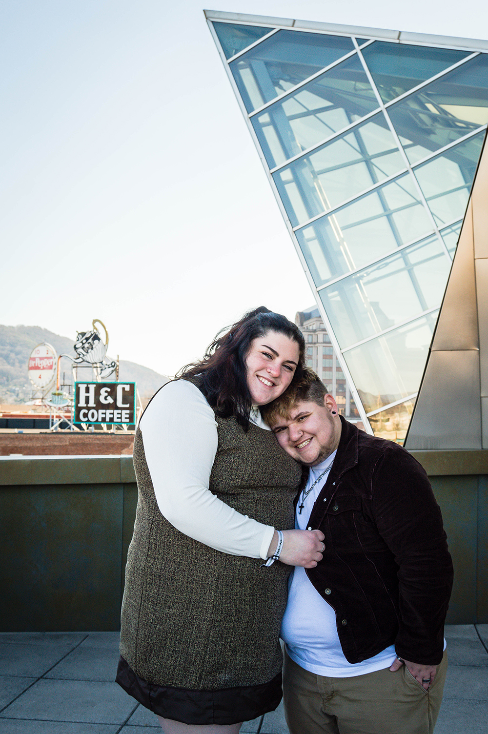 A queer woman rests her cheek on her partner's head and pulls him in closer to her by holding his jacket as he smiles.