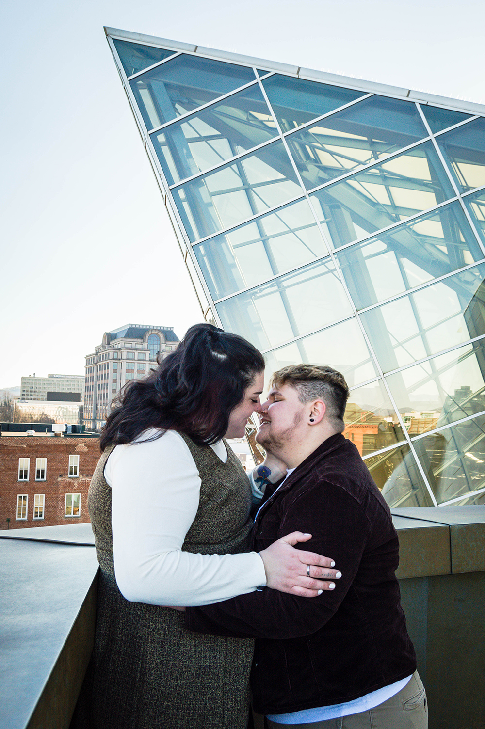 An LGBTQ+ couple go in for a kiss on the balcony of the Taubman Museum of Art for their couples adventure session.