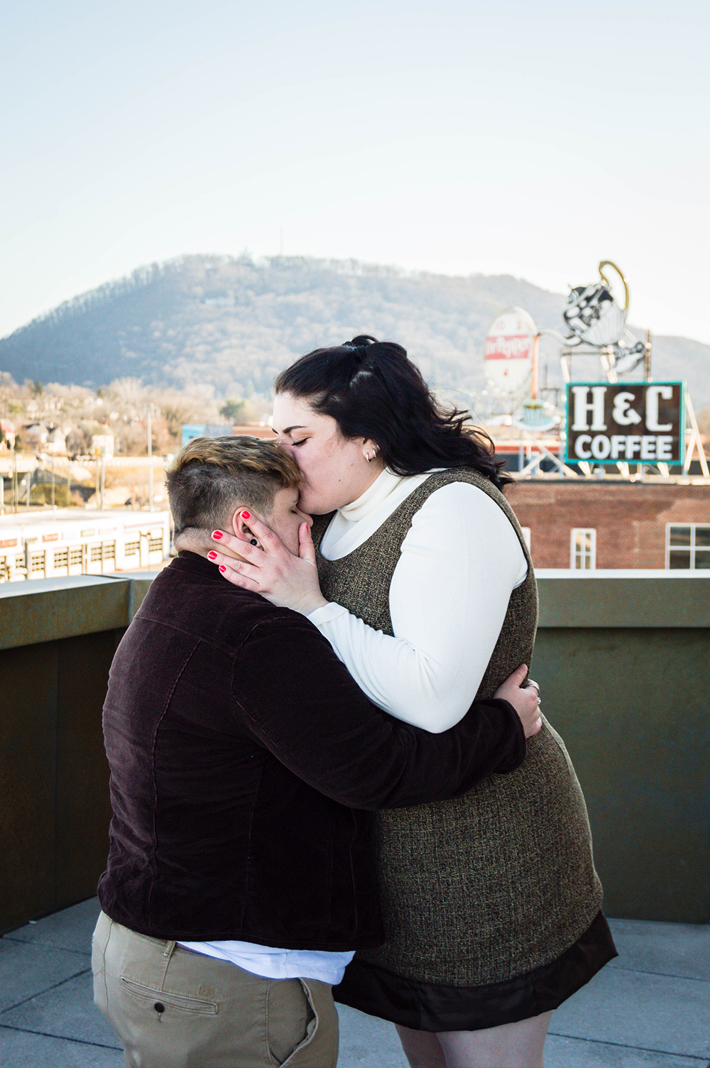 A queer woman kisses the forehead of her trans partner during their couples adventure session on the balcony of the Taubman Museum of Art.