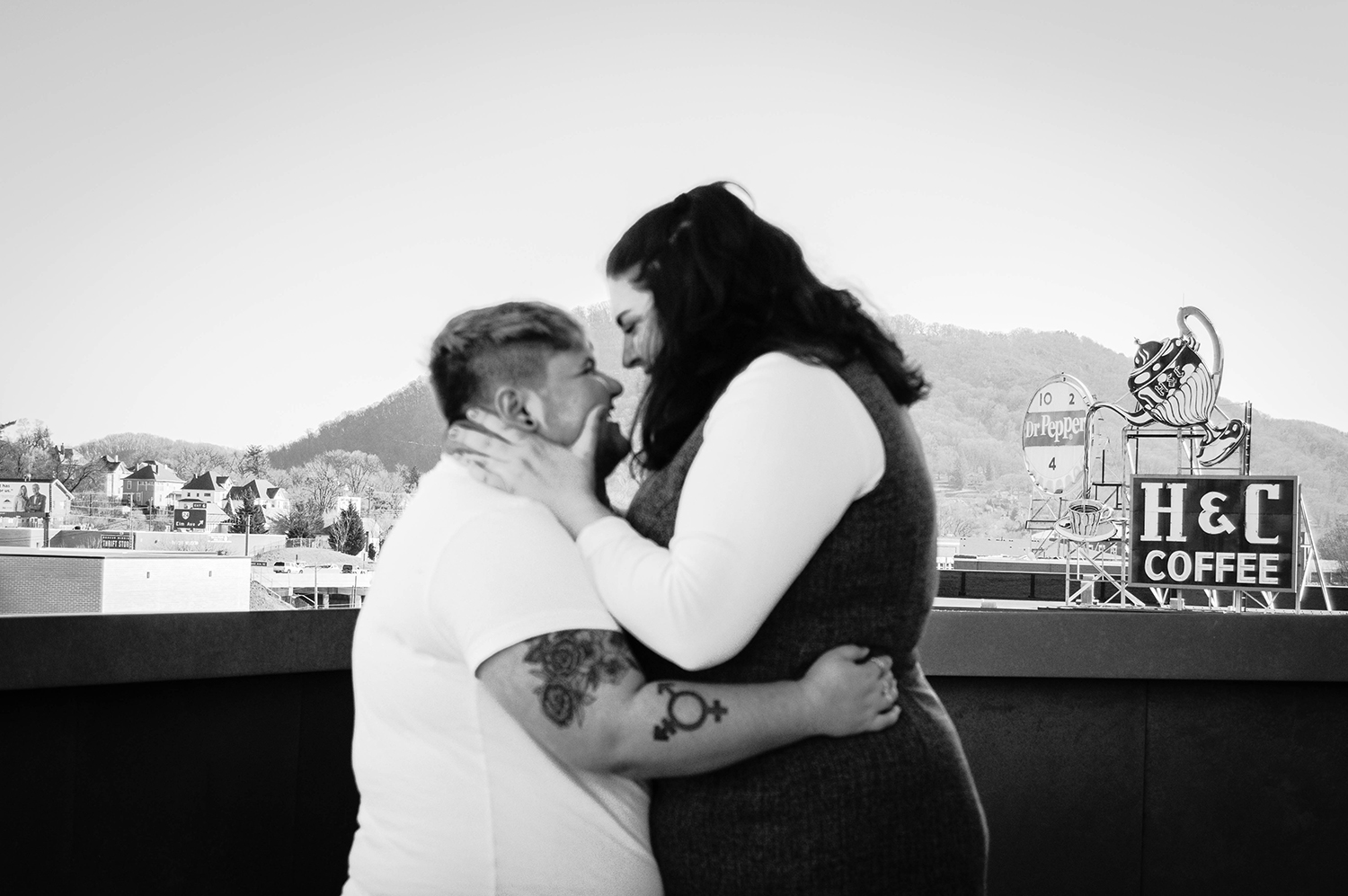 A purposefully blurred out LGBTQ+ couple hold onto one another and smile widely at one another. The background is purposefully focused to reveal the surrounding urban landscape of Roanoke, Virginia, with the iconic H&C Coffee sign and Dr. Pepper sign behind them. 