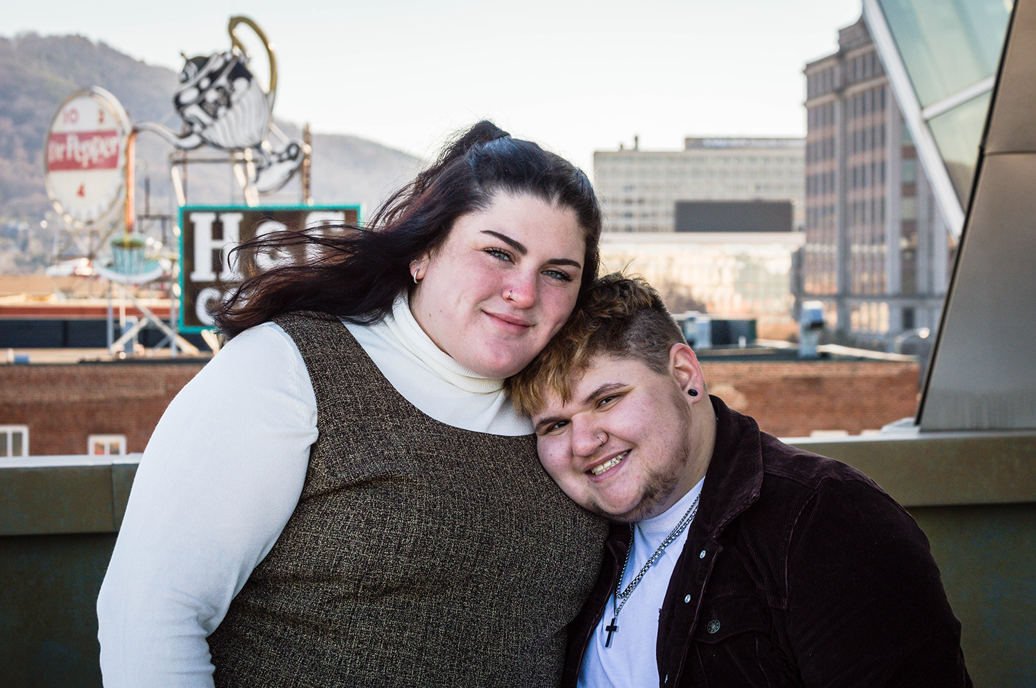 An LGBTQ+ couple stand close to one another and smile for a photo on the Taubman Museum of Art second-floor balcony, which oversees Downtown Roanoke.