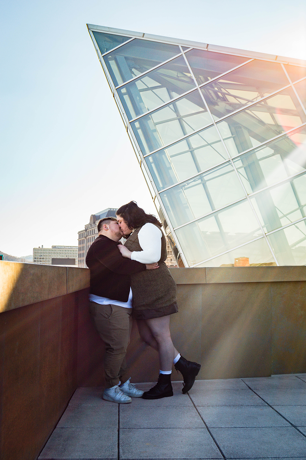 An LGBTQ+ couple kisses one another on the balcony of the Taubman Museum of Art for their couples adventure session as the sun peaks behind part of the museum's glass and steel architecture. 