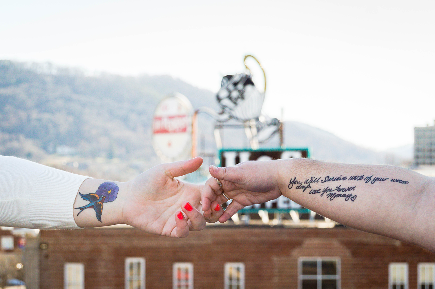 Two hands reach out for one another. On the left side of the image, a person's arm extends and shows a colorful memorial tattoo featuring a bird flying into a flower. On the right side of the photo, another person's arm extends and shows a written tattoo in black ink that reads "you will survive 100% of your worst days. Love you forever. Mommy." with a hand-drawn heart.