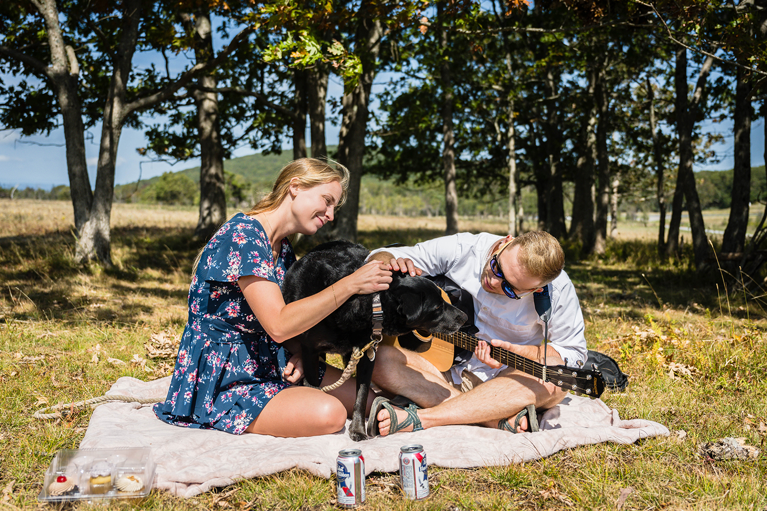 A couple stops what they're doing to pet their dog that has walked in between them as they sit on a picnic blanket on their elopement day.