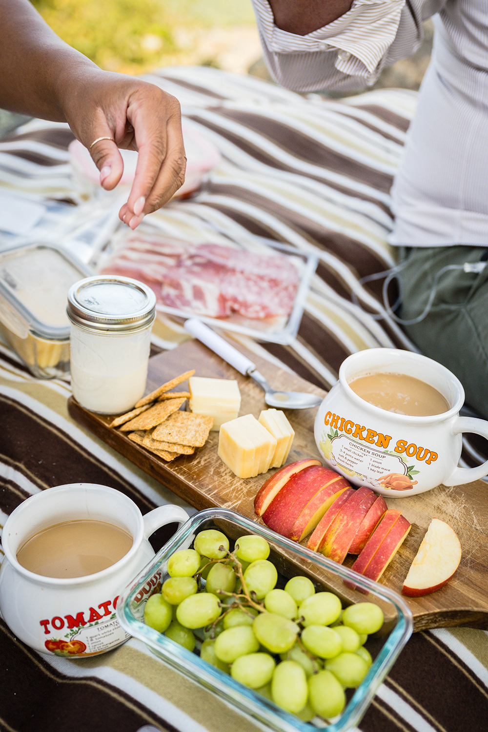 A close up shot of a breakfast charcuterie board with crackers, cheese, meat, and grapes as well as two mugs full of coffee and a mason jar with creamer. One of the marrier's reaches to grab a grape. 