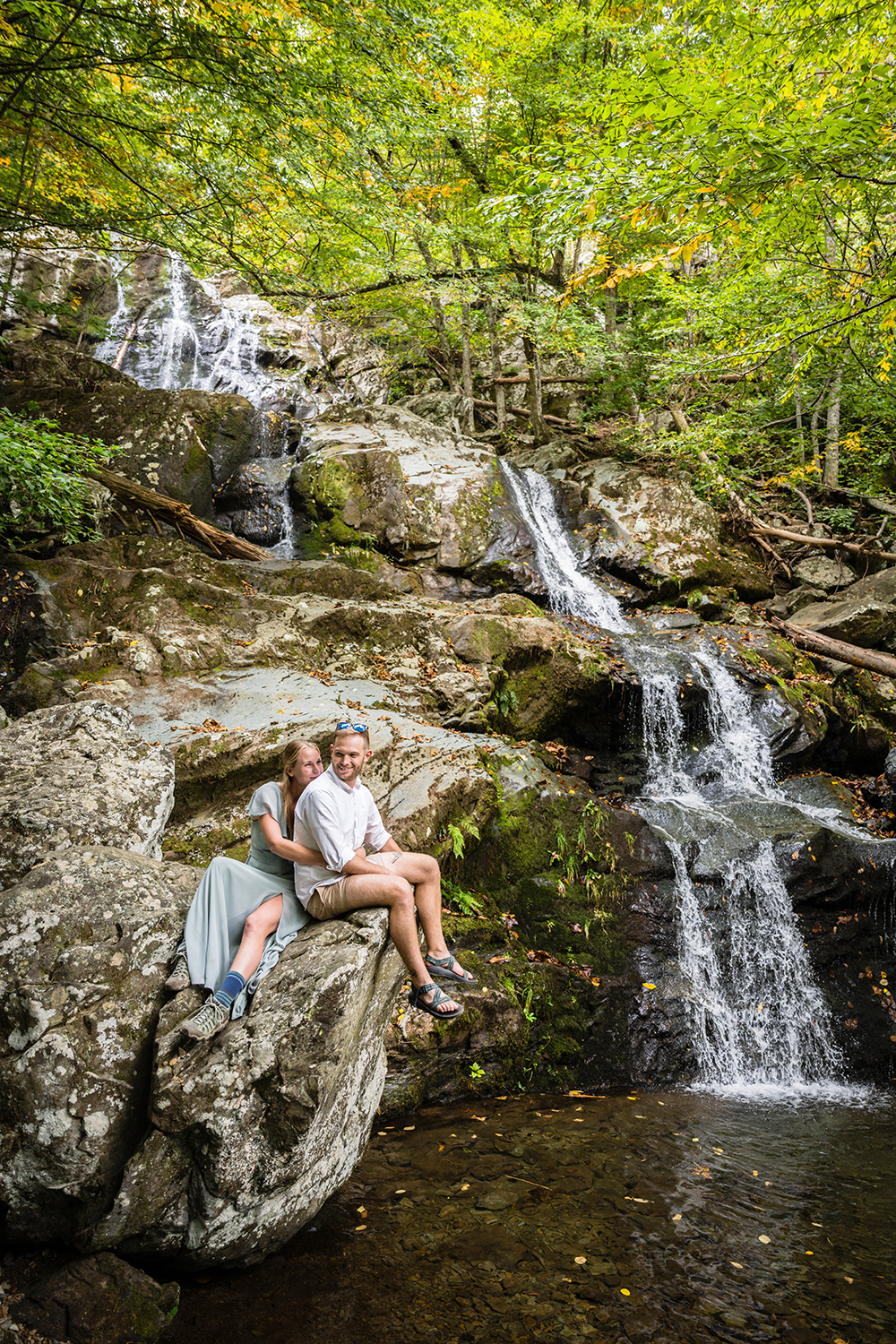 A couple on their elopement day sit on a large rock with a waterfall running behind them. 