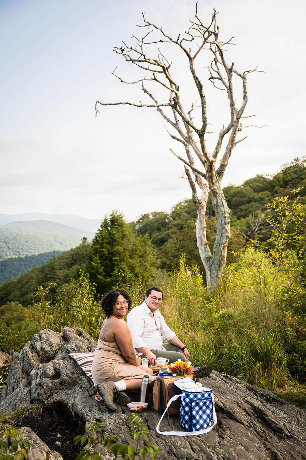 A wedding couple relaxes on a rock, surrounded by nature, with a large tree in the background at Jewel Hollow Overlook in Shenandoah National Park.