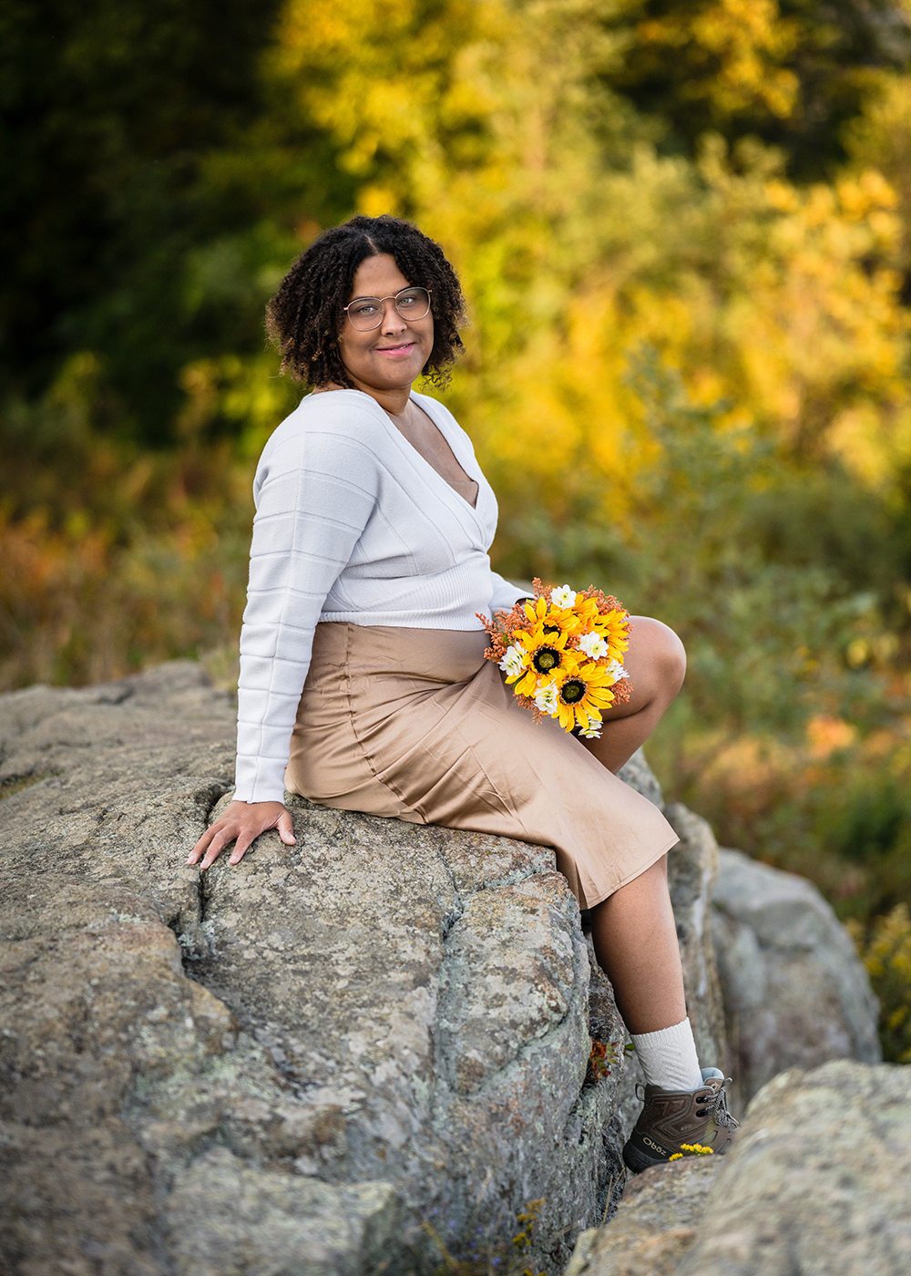 A marrier sits on a large rock holding her bouquet in her lap and letting her feet dangle off the side of the rock at Timber Hollow Overlook in Shenandoah National Park.