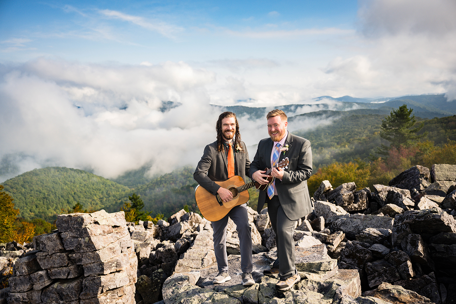 Two queer marriers stand atop rock terrain, each holding an instrument, enjoying the scenic view and the moment together at Blackrock Summit in Shenandoah National Park.