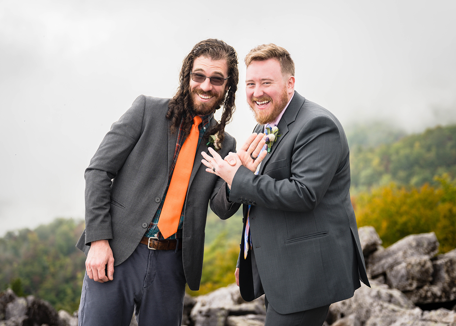 Two marriers in suits pose outdoors with playful expressions. The marrier on the left has long hair and glasses and the marrier on the right has short hair and a beard. Trees and rocks are visible in the background.