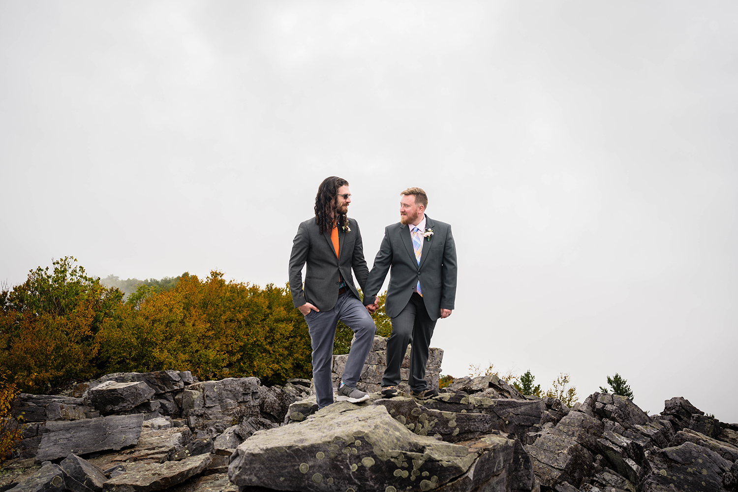 Two gay marriers stand atop rocky mountain terrain with a foggy sky and vibrant orange and green trees in the background at Blackrock Summit in Shenandoah National Park.