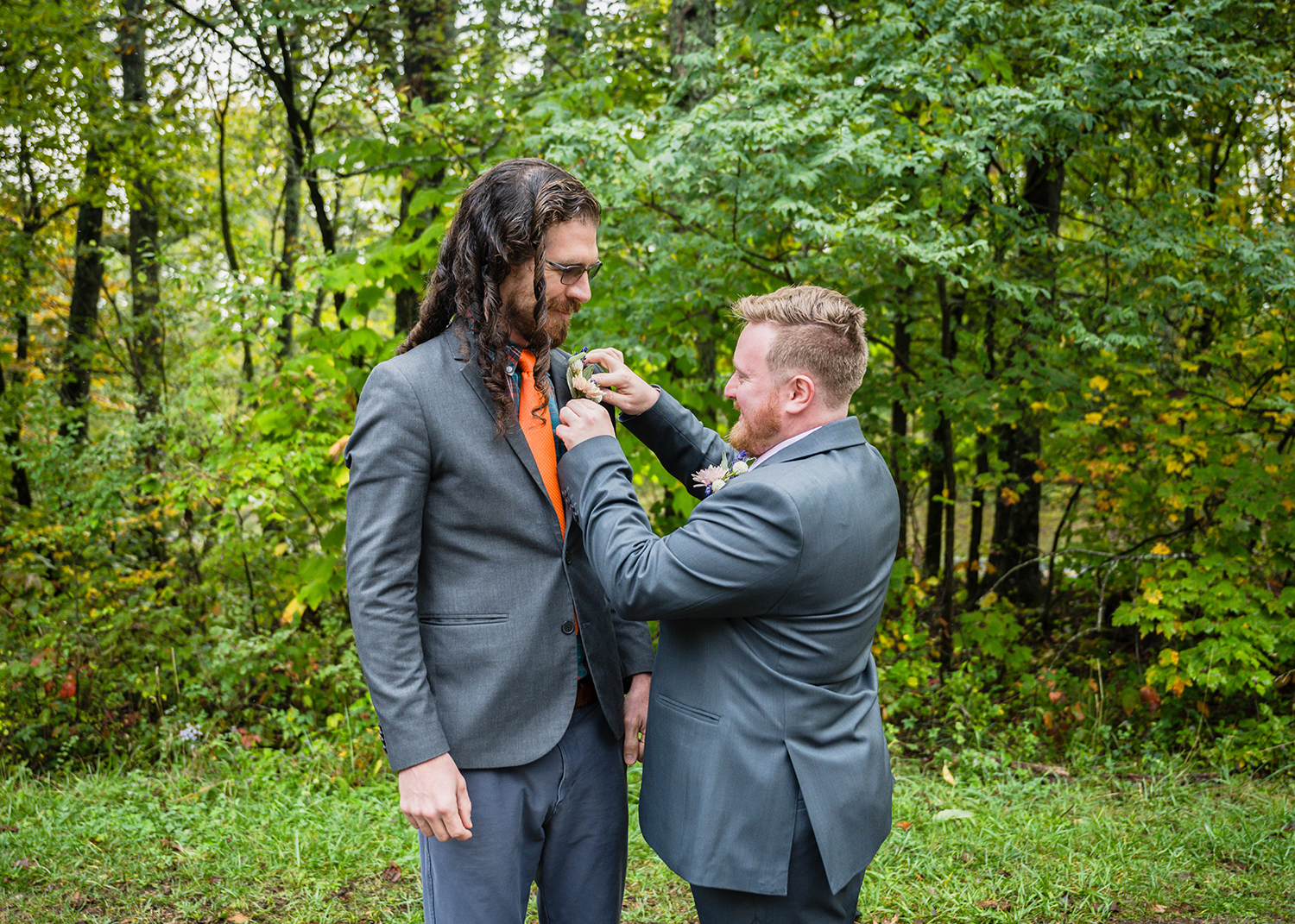 A gay marrier helps their partner pin a boutonniere on their lapel in the forest.