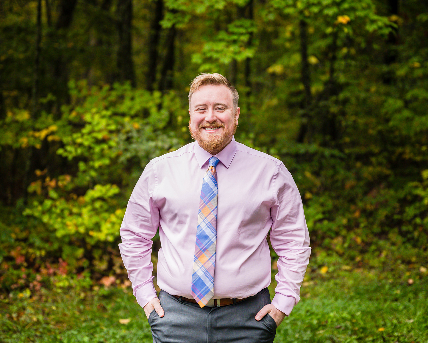 A queer marrier smiles widely with their hands in their pant pockets while standing in the forest for a photo.