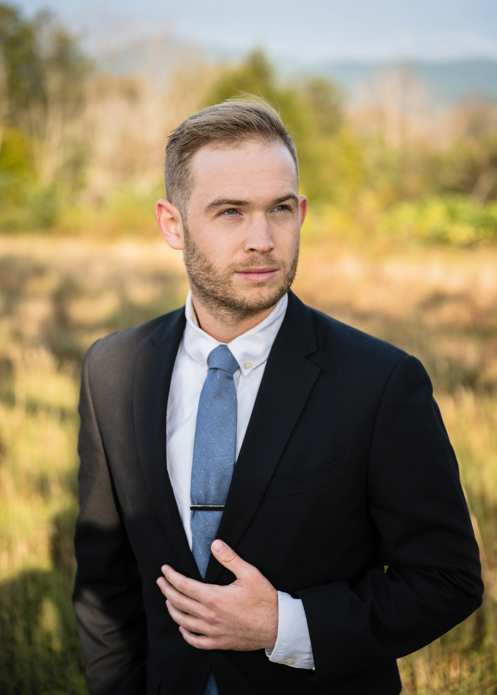 A marrier wearing a suit rests their hand on their stomach and looks off in the distance with a more serious expression on his face. 